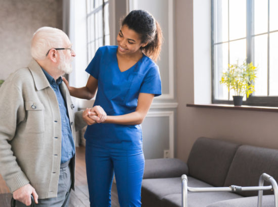 Young Caring African Nurse Helping Senior Old Elderly Man Grandfather Walk . Disable Old Man Trying To Walk With The Assistance Aid Of Female Doctor.