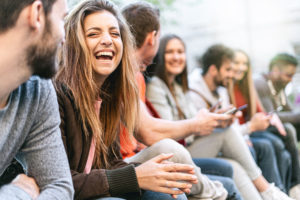 Group Of Trendy Young People Chatting Together Sitting On A Bench Outdoors. Students Having Fun Together. Focus On A Blonde Girl Smiling With Open Mouth