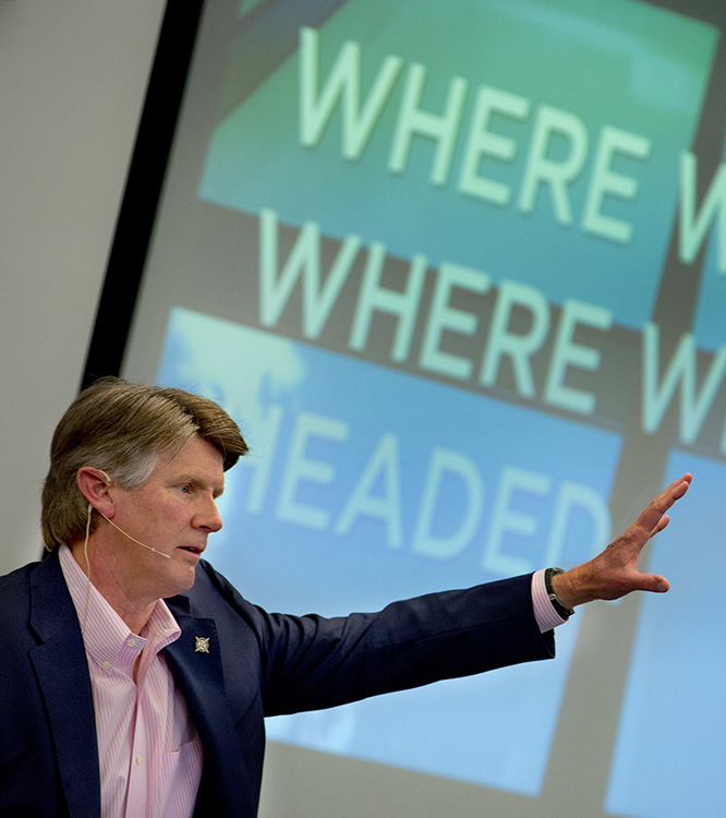 President Williams in front of a projector screen waving his hand during his "See 2020" presentation