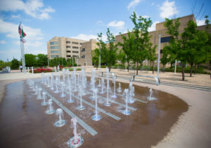 Photo of the Library Courtyard with the water fountains in the front, trees and tables in the middle, and library in the back of the photo.