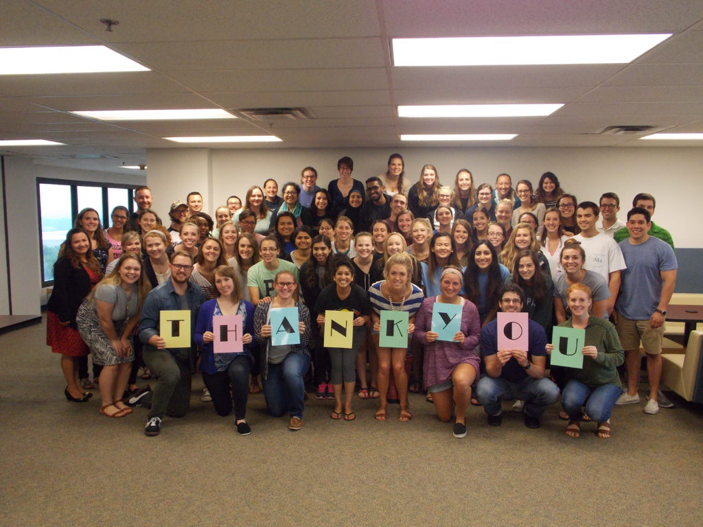 The image shows a large group of people, likely students or a cohort, gathered together in a room. They are all smiling and looking at the camera. In the front row, a few individuals are holding colorful signs that spell out the word "THANK YOU." The group appears to be in a classroom or office environment, with fluorescent lighting and large windows visible in the background.