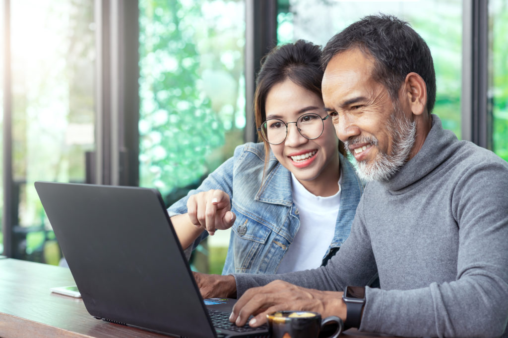 Attractive Mature Asian Man With White Stylish Short Beard Looking At Laptop Computer With Teenage Eye Glasses Hipster Woman In Cafe. Teaching Internet Online Or Wifi Technology In Older Man Concept.